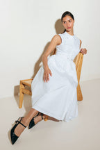 Woman in a white dress sitting on a wooden chair against a plain background