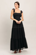 A solid black woven maxi dress featuring straight neckline with lace edge, waist yoke, tiered skirt, bare back with tie closure