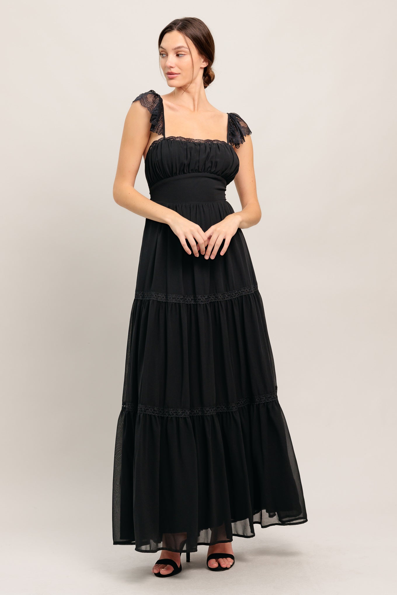 A solid black woven maxi dress featuring straight neckline with lace edge, waist yoke, tiered skirt, bare back with tie closure