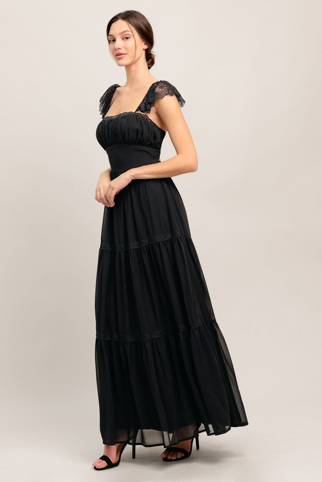 A solid black woven maxi dress featuring straight neckline with lace edge, waist yoke, tiered skirt, bare back with tie closure