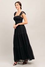 A solid black woven maxi dress featuring straight neckline with lace edge, waist yoke, tiered skirt, bare back with tie closure