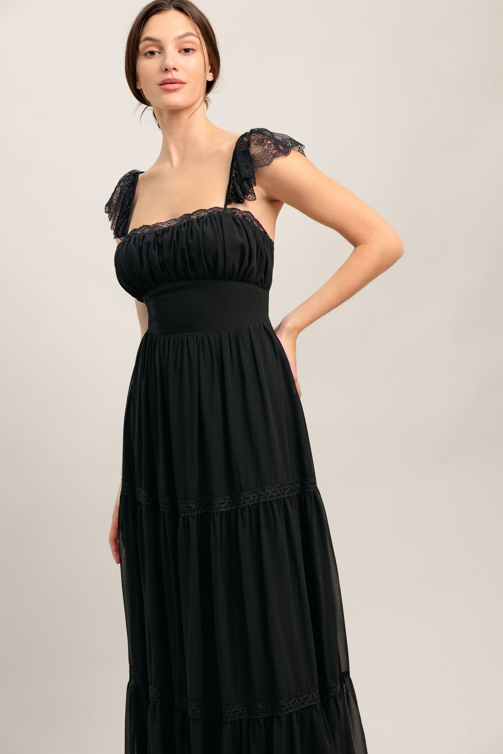A solid black woven maxi dress featuring straight neckline with lace edge, waist yoke, tiered skirt, bare back with tie closure