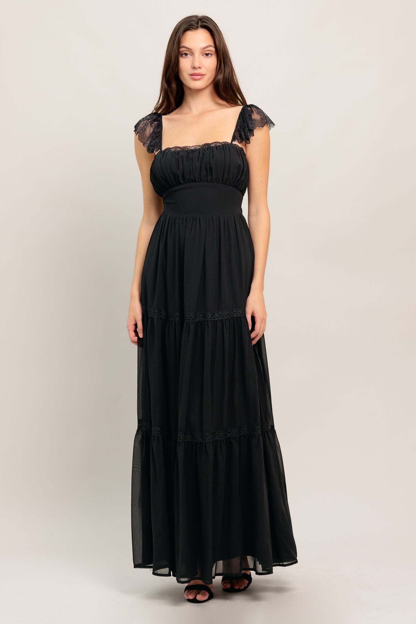 A solid black woven maxi dress featuring straight neckline with lace edge, waist yoke, tiered skirt, bare back with tie closure
