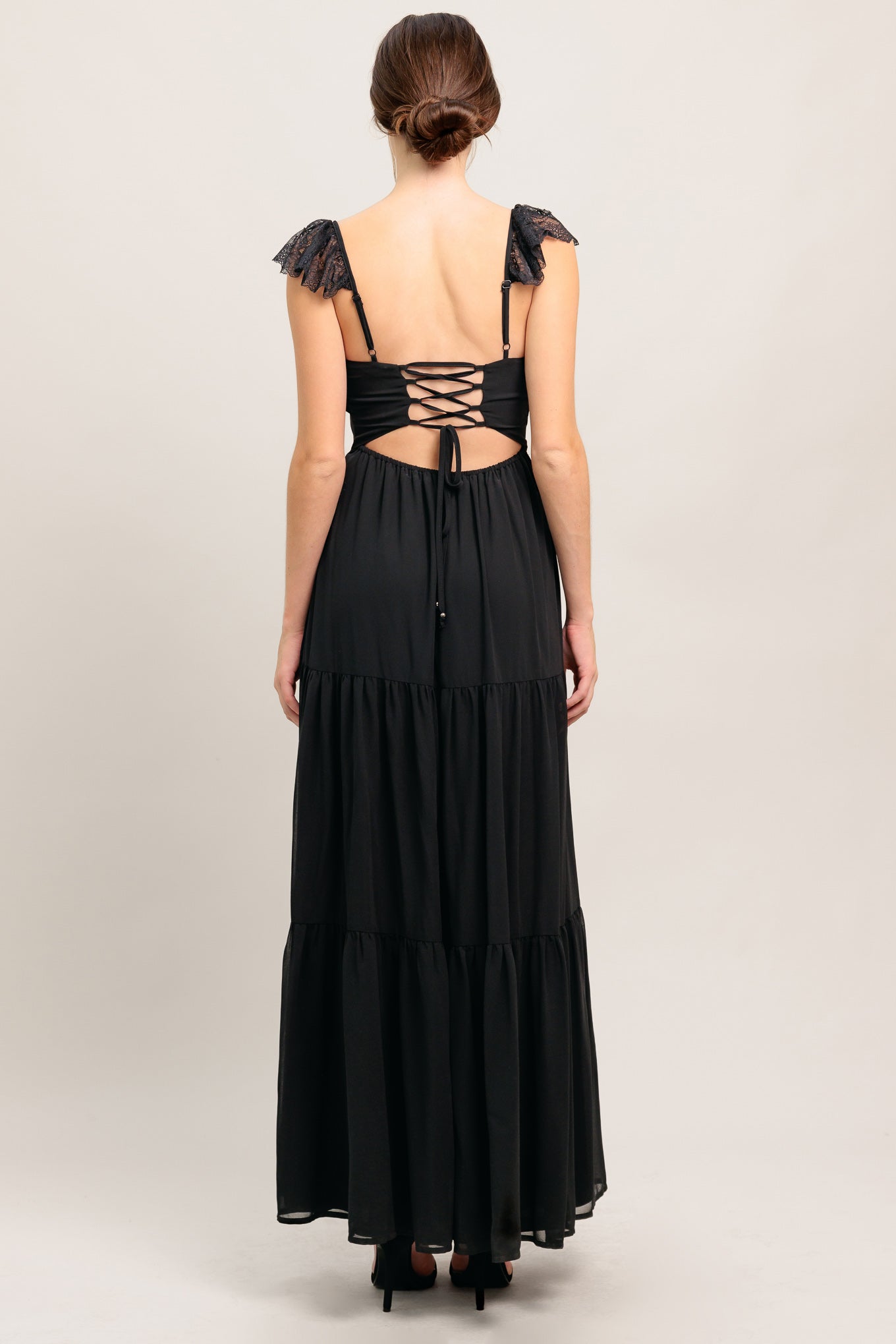 A solid black woven maxi dress featuring straight neckline with lace edge, waist yoke, tiered skirt, bare back with tie closure