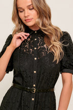 Woman wearing a black lace dress with gold buttons on a plain background